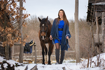 Image showing A girl in a blue dress walks with a horse through the winter forest and old wooden ruins