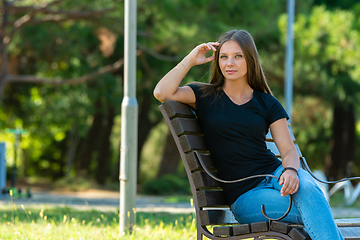 Image showing A beautiful girl in casual clothes sits on a bench in a beautiful green park, the girl looks into the frame