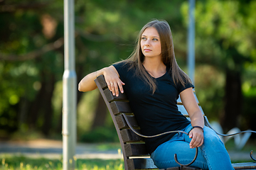 Image showing A beautiful girl in casual clothes sits on a bench in a beautiful green park