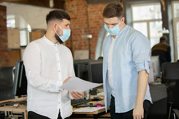 Image showing Young caucasian colleagues working together in a office using modern devices and gadgets during quarantine. Wearing protective face masks