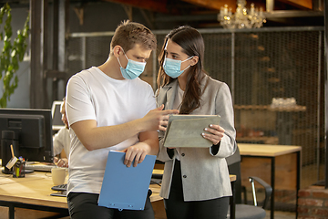 Image showing Young caucasian colleagues working together in a office using modern devices and gadgets during quarantine. Wearing protective face masks