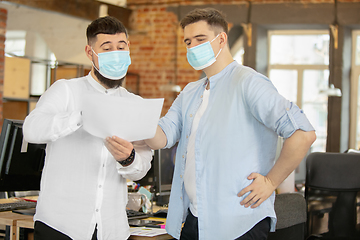 Image showing Young caucasian colleagues working together in a office using modern devices and gadgets during quarantine. Wearing protective face masks