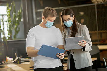 Image showing Young caucasian colleagues working together in a office using modern devices and gadgets during quarantine. Wearing protective face masks