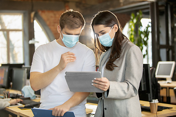 Image showing Young caucasian colleagues working together in a office using modern devices and gadgets during quarantine. Wearing protective face masks