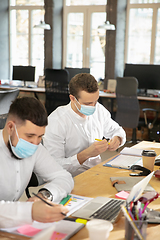 Image showing Young caucasian colleagues working together in a office using modern devices and gadgets during quarantine. Wearing protective face masks