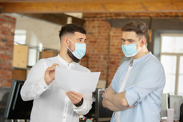 Image showing Young caucasian colleagues working together in a office using modern devices and gadgets during quarantine. Wearing protective face masks
