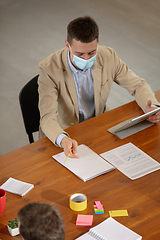 Image showing High angle view colleagues working together in face masks during quarantine in a office using modern devices and gadgets during creative meeting