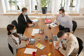 Image showing High angle view colleagues working together in face masks during quarantine in a office using modern devices and gadgets during creative meeting