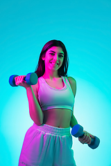 Image showing Brunette beautiful woman\'s portrait isolated on blue studio background in multicolored neon light. Training with dumbbells