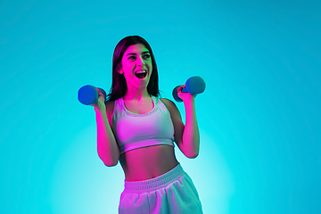 Image showing Brunette beautiful woman\'s portrait isolated on blue studio background in multicolored neon light. Training with dumbbells