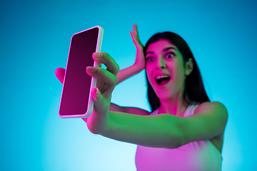 Image showing Brunette beautiful woman\'s portrait isolated on blue studio background in multicolored neon light. Showing blank phone\'s screen