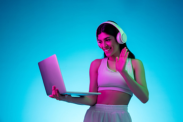 Image showing Brunette beautiful woman\'s portrait isolated on blue studio background in multicolored neon light. Model with headphones and laptop