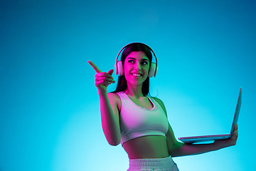 Image showing Brunette beautiful woman\'s portrait isolated on blue studio background in multicolored neon light. Model with headphones and laptop