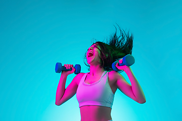 Image showing Brunette beautiful woman\'s portrait isolated on blue studio background in multicolored neon light. Training with dumbbells