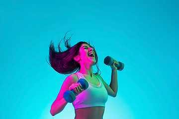 Image showing Brunette beautiful woman\'s portrait isolated on blue studio background in multicolored neon light. Training with dumbbells