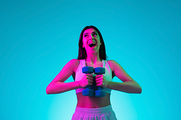 Image showing Brunette beautiful woman\'s portrait isolated on blue studio background in multicolored neon light. Training with dumbbells