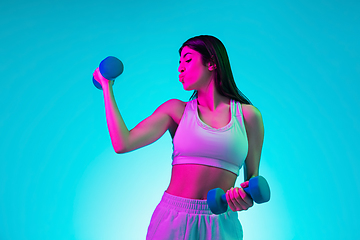 Image showing Brunette beautiful woman\'s portrait isolated on blue studio background in multicolored neon light. Training with dumbbells