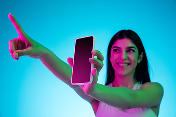 Image showing Brunette beautiful woman\'s portrait isolated on blue studio background in multicolored neon light. Showing blank phone\'s screen