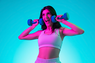 Image showing Brunette beautiful woman\'s portrait isolated on blue studio background in multicolored neon light. Training with dumbbells