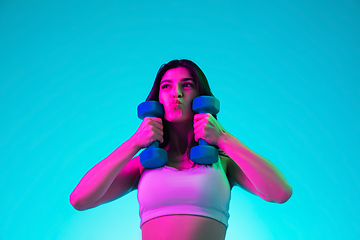 Image showing Brunette beautiful woman\'s portrait isolated on blue studio background in multicolored neon light. Training with dumbbells