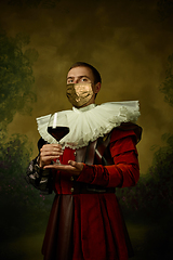 Image showing Portrait of medieval young man in vintage clothing and golden face mask standing on dark background.