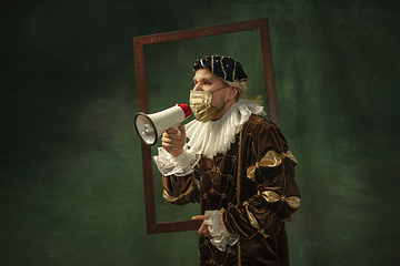 Image showing Portrait of medieval young man in vintage clothing and golden face mask standing on dark background.