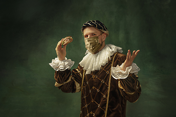 Image showing Portrait of medieval young man in vintage clothing and golden face mask standing on dark background.