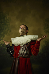 Image showing Portrait of medieval young man in vintage clothing and golden face mask standing on dark background.