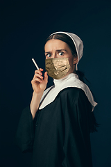 Image showing Medieval young woman as a nun in vintage clothing and golden face mask on dark blue background. Concept of comparison of eras, protection from covid