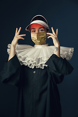 Image showing Medieval young woman as a nun in vintage clothing and golden face mask on dark blue background. Concept of comparison of eras, protection from covid