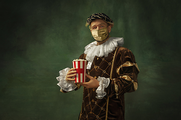 Image showing Portrait of medieval young man in vintage clothing and golden face mask standing on dark background.