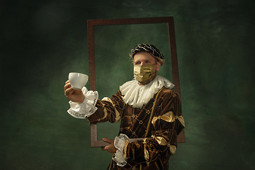 Image showing Portrait of medieval young man in vintage clothing and golden face mask standing on dark background.