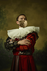 Image showing Portrait of medieval young man in vintage clothing and golden face mask standing on dark background.