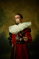 Image showing Portrait of medieval young man in vintage clothing and golden face mask standing on dark background.