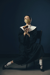 Image showing Medieval young woman as a nun in vintage clothing and golden face mask on dark blue background. Concept of comparison of eras, protection from covid