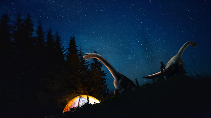 Image showing Dinosaurs huge higth walking through the night forest comes across tourists group in travel tent