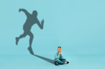 Image showing Childhood and dream about big and famous future. Conceptual image with boy and shadow of fit male runner on blue background