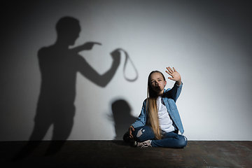 Image showing Domestic violence, abusing. Scared little caucasian girl, victim sitting close to white wall with shadow of angry father\'s threatening on it.