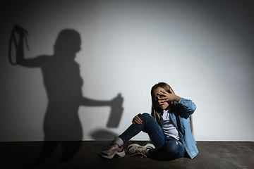 Image showing Domestic violence, abusing. Scared little caucasian girl, victim sitting close to white wall with shadow of angry threatening mother with alcohol addiction.