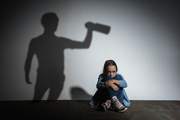 Image showing Domestic violence, abusing. Scared little caucasian girl, victim sitting close to white wall with shadow of angry threatening father with alcohol addiction.