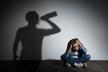 Image showing Domestic violence, abusing. Scared little caucasian girl, victim sitting close to white wall with shadow of angry threatening father with alcohol addiction.