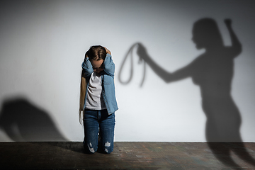 Image showing Domestic violence, abusing. Scared little caucasian girl, victim sitting close to white wall with shadow of angry mother\'s threatening on it.