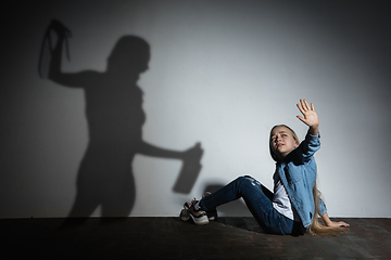 Image showing Domestic violence, abusing. Scared little caucasian girl, victim sitting close to white wall with shadow of angry threatening mother with alcohol addiction.