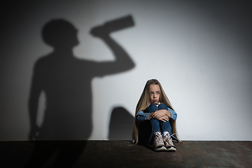 Image showing Domestic violence, abusing. Scared little caucasian girl, victim sitting close to white wall with shadow of angry threatening father with alcohol addiction.