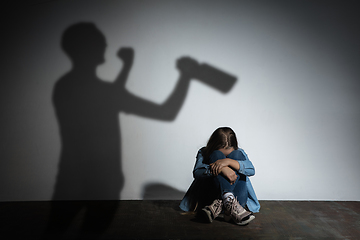 Image showing Domestic violence, abusing. Scared little caucasian girl, victim sitting close to white wall with shadow of angry threatening father with alcohol addiction.