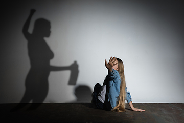 Image showing Domestic violence, abusing. Scared little caucasian girl, victim sitting close to white wall with shadow of angry threatening mother with alcohol addiction.