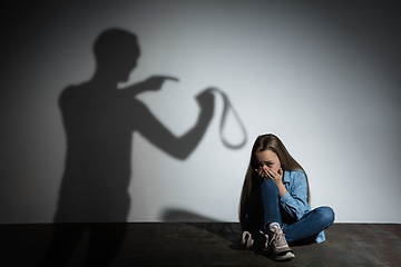 Image showing Domestic violence, abusing. Scared little caucasian girl, victim sitting close to white wall with shadow of angry father\'s threatening on it.