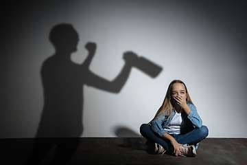 Image showing Domestic violence, abusing. Scared little caucasian girl, victim sitting close to white wall with shadow of angry threatening father with alcohol addiction.