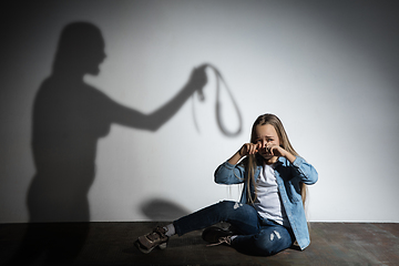 Image showing Domestic violence, abusing. Scared little caucasian girl, victim sitting close to white wall with shadow of angry mother\'s threatening on it.