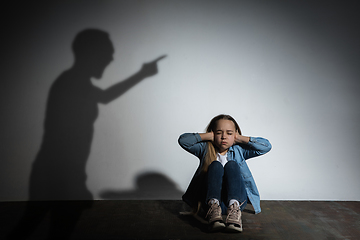 Image showing Domestic violence, abusing. Scared little caucasian girl, victim sitting close to white wall with shadow of angry father\'s threatening on it.